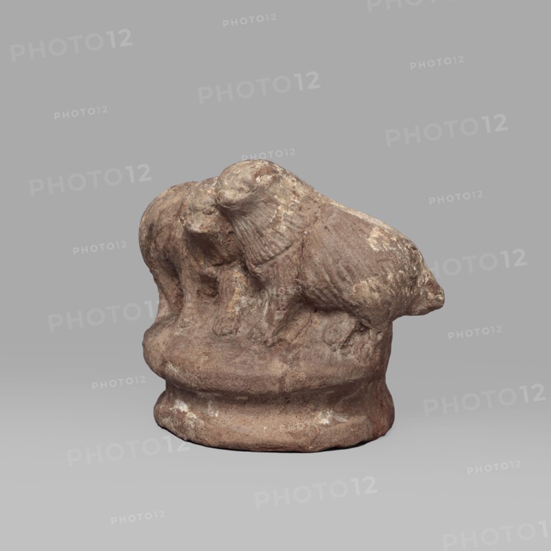 Two dogs, Two dogs shown schematically on a round, profiled base. The dogs are shown standing, with their heads touching. Ptolemaic Period, 332–30 BC; Tell Atrib. Figurine; height 5.3 cm, width 7.1 cm., A small weathered sculpture of two animals standing close together on a rounded base, both showing textured fur and rounded heads; the piece appears rough and eroded with uneven surfaces and small patches of lighter wear. The visible colors are brown, tan, beige, and hints of off-white against a neutral gray background.