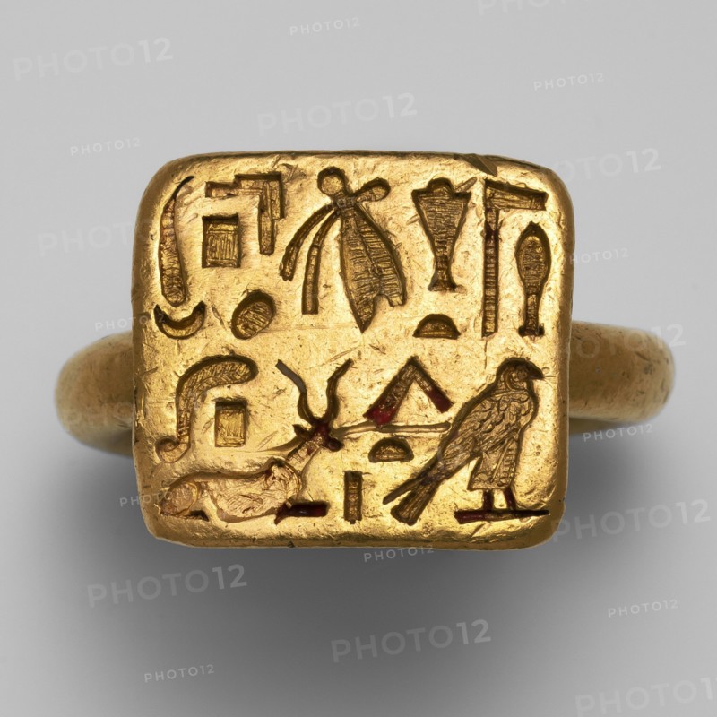 Gold ring of the high priest of the goddess Bastet, Hapimen ca. 664–525 BC. Goldsmithery; gold ring with a rectangular plaque on a circular thick-cylinder hoop. Upper surface of the plaque covered with two rows of hieroglyphic inscription in concave relief. Date: 664–525 BC, Late Period, 26th Dynasty. Possible origin: Leontopolis, possible findspot: Tell el-Moqdam . Subject: Hapimen, Bastet. Dimensions: total length 2.6 cm, width 2.6 cm; eyelet height 1.6 cm, width 1.8 cm; ring thickness 0.4 cm., A gold ring with a square bezel engraved with a group of carved symbols and small pictorial shapes, showing a bird figure, an insect-like motif, a reclining animal shape, and various geometric and curved forms; the ring and bezel are metallic gold in color with small areas of darker brownish or reddish insets within some carved lines, set against a neutral light gray background.