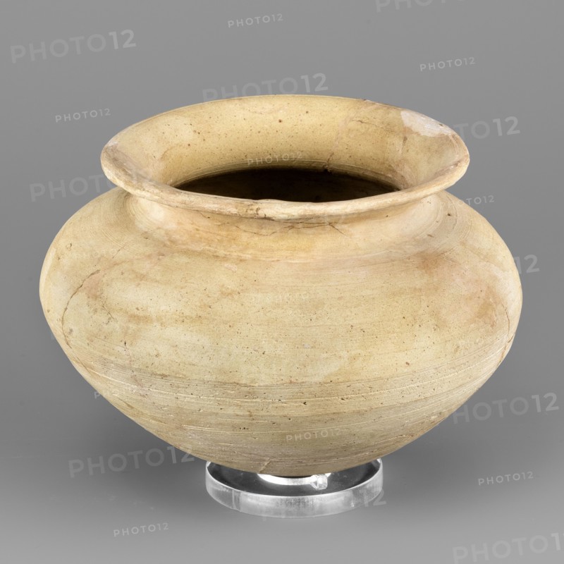 Vessel with an egg-shaped base, unknown Northern Mesopotamian workshop; vessel for oils and ointments, vice-free vessel. Vessel with an egg-shaped base and a bulging, conically inclined belly, transitioning into short, rounded shoulders with a low, inclined neck terminating in a narrow, outwardly curled rim. On the inside, the neck is separated from the rim by a thin engraved line. 1st half of the 3rd millennium BC; Early Jazira III period Early Dynastic III; ca. 2600–ca. 2350 BC. Tell Abu Hafur North-Eastern Syria, Northern Mesopotamia Khabur basin. Vessel; height 9.2 cm, width 13.3 cm, weight 270 g., A rounded, wide-mouthed ceramic vessel sits on a clear circular stand against a plain background; the pot has a bulbous body with a slightly flared rim and visible surface irregularities and faint circular marks from shaping. The visible colors are beige, light tan, cream, pale yellow, and touches of brown, with subtle darker areas inside the opening and slight discolorations and streaks...