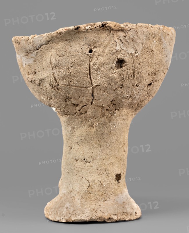 Beaker with engraved decoration, unknown northern Mesopotamian workshop. Hand-made beaker on a thick, cylindrical stem with a poorly defined foot. The bowl is a low form with an irregularly defined rim, glued to the foot. Three schematically engraved human figures on the bowl: the torso with head and legs is a vertical line terminating in a dot at the top and a short arc opening downward at the bottom; the arms, extended upward, are marked by an engraved horizontal line curved at both ends that intersects the torso slightly below its midpoint. 1st half of the 3rd millennium BC; Early Jazira III period Early Bronze Period; Early Dynastic III period; ca. 2600–ca. 2350 BC. Tell Abu Hafur northeastern Syria, Khabur basin; Northern Mesopotamia Khabur basin. Vessel; weight 148 g., A weathered, textured cup-like vessel with a wide bowl atop a narrow stem and a flared base, shown against a plain gray background. The object has an uneven, rough surface with visible cracks, pits, and small holes...