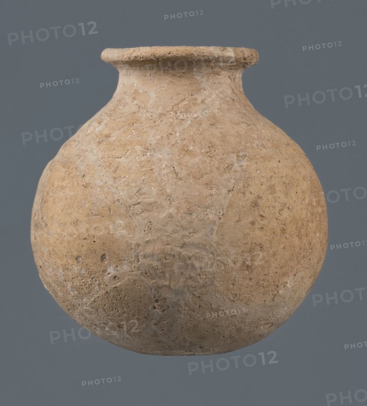Globular vessel, unknown Northern Mesopotamian workshop, Early Jazira III period ca. 2600–ca. 2350 BC Early Dynastic III period. A globular vessel whose body rises to three-quarters height into sloping arms, from which a low narrow neck arises, terminating in a thickened, flared rim. The transition from body to arms is emphasized by a shallow, wide horizontal groove. Circa 2600–ca. 2350 BC. Origin: Northern Mesopotamia Khabur basin, Tell Jassa el Gharbi, northeastern Syria. Function: vessel for oils and ointments; vice-free vessel. Material/type: vessel; height 10 cm, width 9.7 cm., A rounded, bulbous clay vessel with a short neck and flared rim sits centered against a solid muted gray background; the vessel surface is rough and textured with an uneven, weathered appearance showing a mix of beige, tan, light brown, and pale gray tones.