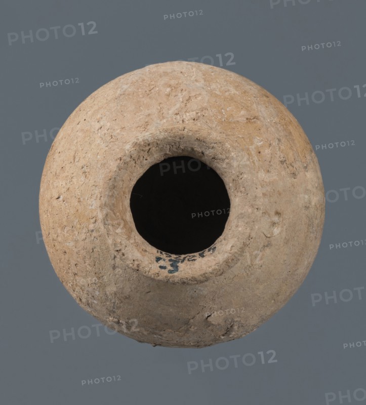 A globular vessel, likely from a Northern Mesopotamian workshop, Early Jazira III / Early Dynastic III period ca. 2600–ca. 2350 BC. The body rises to about 3/4 height into sloping shoulders from which a low, narrow neck emerges, terminating in a thickened, flared rim; the transition from body to shoulders is emphasized by a shallow, wide horizontal groove. Made for oils and ointments; vice-free ceramic. Origin: Al-Jazira Khabur basin, Northern Mesopotamia, Tell Jassa el Gharbi Northeastern Syria. Dimensions: height 10 cm; width 9.7 cm., A round, rough-textured clay vessel with an open circular mouth viewed from above, showing a dark empty interior and uneven, weathered surfaces with small pits and chips; the vessel is in shades of beige, tan, light brown, gray, black inside the opening, and the background is a muted slate gray.