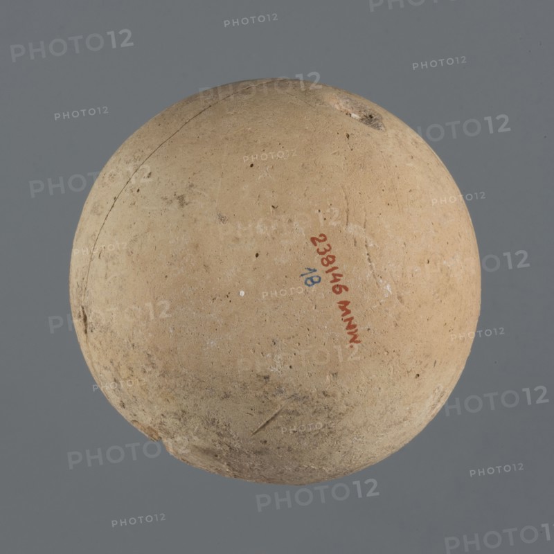 Globular vessel, unknown Northern Mesopotamian workshop Khaburu Basin, Early Jazira III period ca. 2600–ca. 2350 BC, vessels for oils and ointments, clay vessel, Early Dynastic III period ca. 2600–ca. 2350 BC, Al-Jazira Syria; Iraq; Mesopotamia, globular vessel with a spherical belly that at 3/4 height merges into short, sloping shoulders with a narrow, slightly flared neck; part of the neck and spout missing. c. 2600–2350 BC; Northern Mesopotamia Khabur basin, Tell Jassa el Gharbi north-eastern Syria. Vessel; height 13 cm, width 10.9 cm., A roughly spherical object with a textured, worn surface in tan and beige tones sits against a flat gray background, showing small pits, scratches, and a few darker spots and smudges; there is a small area of chipped or rougher material near the top and a slightly darker, dirtier band along the lower edge, with subtle variations of light and shadow across the surface.