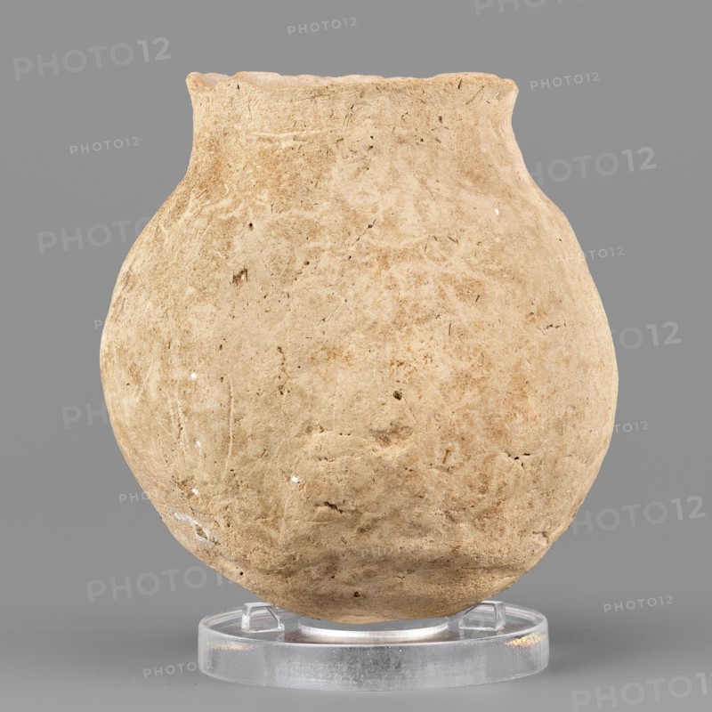 Spherical vessel, unknown Northern Mesopotamian workshop, viseless, hand-made miniature vessel with a rounded bottom and a low neck transitioning into a slightly tilted and undulating short rim with a pointed tip; irregular, wide vertical grooves on the body. 1st half of the 3rd millennium BC; Early Jazira III period ca. 2600–ca. 2350 BC. Tell Jassa el Gharbi North-Eastern Syria; Northern Mesopotamia Khabur basin. Vessel; height 7.9 cm, width 7.1 cm, weight 176 g., A single rounded vessel with a rough, textured surface sits upright on a clear circular stand against a solid gray background; the vessel is an uneven beige to light tan color with small darker flecks and faint variations in tone across its body, showing irregular pits and scratches in the clay and a slightly wavy rim, while the clear stand appears translucent with subtle reflections.