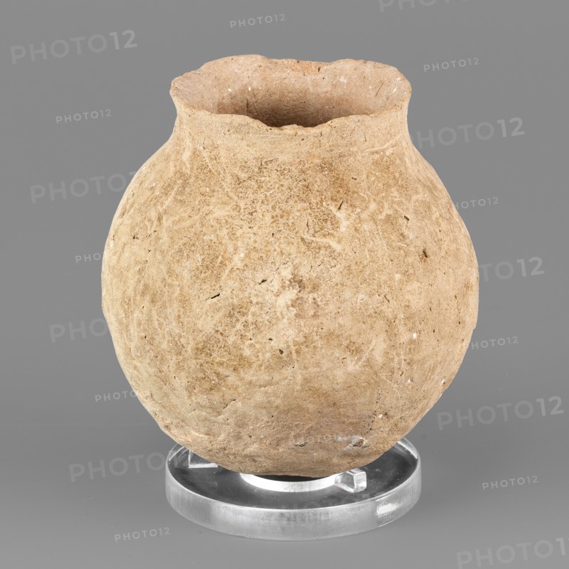 Spherical vessel, unknown Northern Mesopotamian workshop; hand-made miniature spherical vessel with a rounded bottom and a low neck transitioning into a slightly tilted, undulating short rim with a pointed tip; irregular, wide vertical grooves on the body; viseless, hand-made miniature vessel; Near Eastern pottery; Eastern Gaulish Scene style; 1st half of the 3rd millennium BC, Early Jazira III period ca. 2600–ca. 2350 BC; Tell Jassa el Gharbi North-Eastern Syria, Northern Mesopotamia Khabur basin; vessel; height 7.9 cm, width 7.1 cm, weight 176 g., A rounded, textured clay vessel with an uneven rim sits on a clear circular stand against a flat background; the vessel shows a rough, earthy surface with small pits and irregularities and appears in shades of tan, beige, and light brown, while the stand is translucent and slightly reflective, and the background is a uniform medium gray.