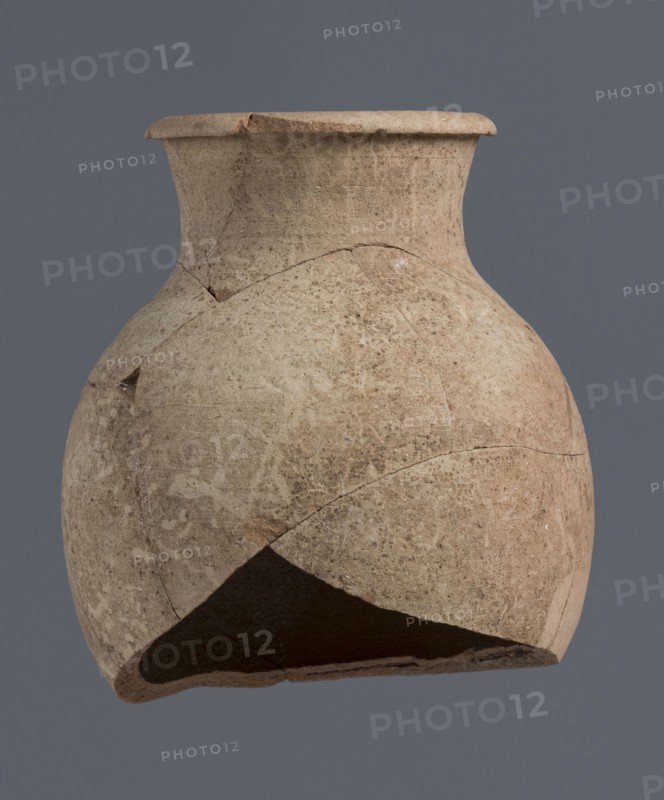 Fragment of a bottle upper 3/4, unknown Northern Mesopotamian workshop, Early Jazira III period ca. 2600–ca. 2350 BC Early Dynastic III period. The upper 3/4 of a vice-free bottle with a slender ovoid belly and a high neck terminating in a thickened, outwardly turned-out rim of triangular cross-section. Clay vessel bottle. Provenance: Tell Jassa el Gharbi north-eastern Syria, Northern Mesopotamia Khabur basin. Height 18 cm; width 16 cm., A weathered, rounded clay vessel with a flared rim and a narrow neck sits against a plain gray background, showing a surface of beige and light brown tones with patches of darker brown and pale speckling; the vessel displays several visible cracks and areas where pieces are missing, revealing an inner, darker interior, and the exterior texture appears rough and porous with irregular shading and subtle worn marks.