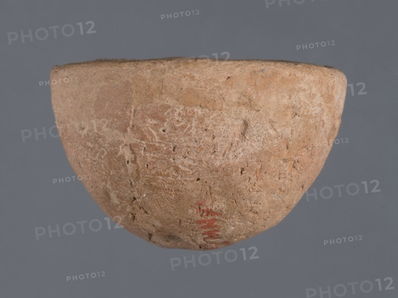 Miniature bowl, unknown Northern Mesopotamian workshop, Early Jazira III period ca. 2600–ca. 2350 BC. Clay; miniature vessel with a narrow rounded missing bottom and a straight rim; arched walls terminating in a straight rim. Early Dynastic III period ca. 2600–ca. 2350 BC. Origin: Northern Mesopotamia Khabur basin, Tell Jassa el Gharbi North-Eastern Syria. Height 3 cm; width 5 cm., A roughly hemispherical bowl with a textured, worn surface in shades of tan, beige, and light brown is centered against a solid gray background; the bowl shows areas of roughness and small pits, with subtle variations of cream and ochre on its exterior and a small area of muted red near the lower rim.