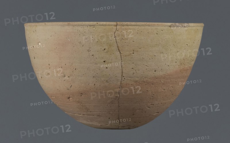 Bowl, unknown Northern Mesopotamian workshop. Early Jazira III period ca. 2600–ca. 2350 BC / Early Dynastic III period. Small round flat-bottomed bowl: walls tilted in the lower half, straight in the upper half, ending with a low, slightly distinct, slightly thickened and rounded rim. Pinkish burn mark visible on the lower part from contact with another bowl. Material: clay. Origin: Northern Mesopotamia Al-Jazira/Khabur Basin, found at Tell Jassa el Gharbi Northeastern Syria. Function: vessel/table bowl. Dimensions: height 7.7 cm; width 12.3 cm; weight 170 g., A rounded ceramic bowl shown against a solid dark gray background, with a visible vertical crack running from rim to base and a surface pitted with small holes and irregularities; the bowl displays a gradient of colors including beige, tan, light brown, pale pinkish tones, and hints of yellow, with subtle horizontal striations and areas of darker brown near the lower portion.