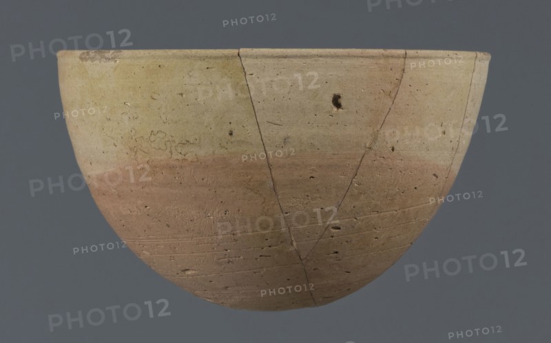 Bowl, unknown Northern Mesopotamian workshop. Early Jazira III period ca. 2600–ca. 2350 BC / Early Dynastic III period ca. 2600–ca. 2350 BC. Clay vessel, table vessel; vice-free. Bowl with a small round flat bottom; walls tilted in the lower half, straight in the upper half, ending with a low, slightly distinct, slightly thickened and rounded rim. Pinkish burn mark in the lower part from contact with the bowl in which it was placed. Ca. 2600–2350 BC. Findspot: Tell Jassa el Gharbi Northeastern Syria, Northern Mesopotamia Khabur Basin. Height 7.7 cm; width 12.3 cm; weight 170 g., A shallow bowl shape with a rounded base and flared rim sits against a solid gray background; the object shows a smooth, worn surface with a gradient of muted colors including beige, pale yellow, light tan, and a washed reddish-brown band across the lower half, and there are visible hairline cracks, small pitted holes, and irregular surface marks across the exterior.