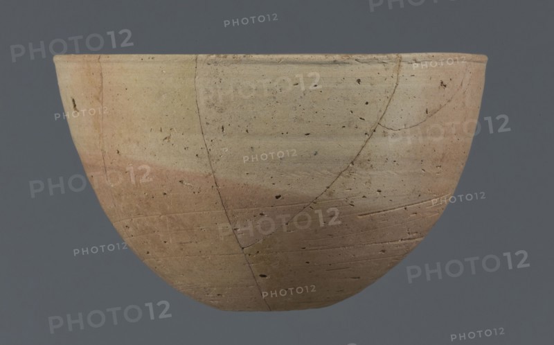 Bowl, unknown Northern Mesopotamian workshop. Bowl with a small round flat bottom. The bowl walls are tilted in the lower half, straight in the upper half, ending with a low, slightly distinct, slightly thickened and rounded rim. In the lower part of the bowl, a pinkish burn mark is visible, left by the bowl in which it was placed. Clay vessel, table vessel; vice-free vessel. Early Jazira III period ca. 2600–ca. 2350 BC / Early Dynastic III period ca. 2600–ca. 2350 BC. Al-Jazira Northern Mesopotamia; Khabur Basin, Tell Jassa el Gharbi Northeastern Syria, ca. 2600–ca. 2350 BC. Height 7.7 cm; width 12.3 cm; weight 170 g., A shallow rounded bowl viewed from the side against a gray background, showing a weathered surface with a gradient of beige to light brown and hints of pink, small dark speckles and pits across the surface, several visible hairline cracks and lines forming irregular patterns, and subtle horizontal striations and texture variations on the clay.