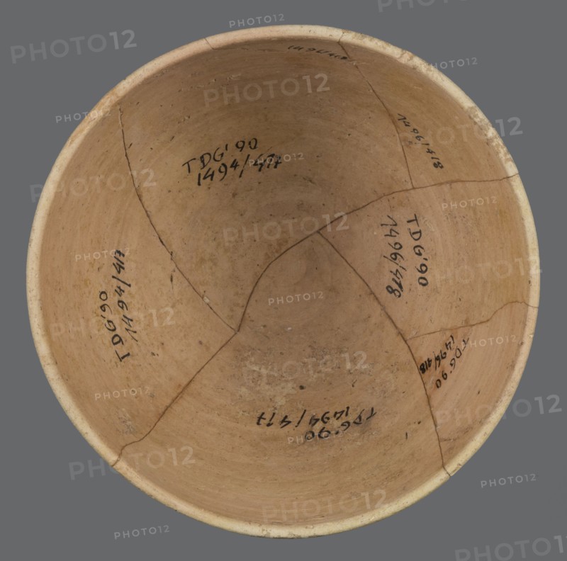 Bowl, unknown Northern Mesopotamian workshop, bowl with a small round flat bottom. The bowl walls are tilted in the lower half, straight in the upper half, ending with a low, slightly distinct, slightly thickened and rounded rim. In the lower part of the bowl, a pinkish burn mark is visible, left by the bowl in which it was placed. Clay vessel, table vessel, vice-free, ca. 2600–2350 BC; Early Jazira III / Early Dynastic III period. Found at Tell Jassa el Gharbi Northeastern Syria; origin Northern Mesopotamia Khabur Basin. Height 7.7 cm, width 12.3 cm, weight 170 g., A round, shallow ceramic bowl viewed from above with an interior surface in shades of tan and beige showing a slightly darker central area and lighter rim; the bowl has visible cracks and a few repaired seams forming a patchwork of segments, with some small spots of discoloration and surface irregularities; the background is a flat gray.