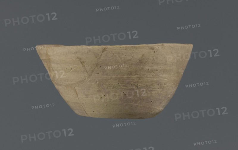 Bowl, unknown Northern Mesopotamian workshop, Early Jazira III period ca. 2600–ca. 2350 BC. Clay table vessel with a flat round bottom showing traces of trimming. Straight, flaring conical walls terminating in a straight, non-distinctive rim; the whole is asymmetrical. Circa 2600–2350 BC. Tell Jassa el Gharbi north-eastern Syria, Northern Mesopotamia Khabur Basin., A shallow, wide bowl with an uneven rim and a rounded base, showing a rough, textured surface with visible tool marks and small pits; the bowl appears in shades of beige and light brown with subtle variations and patches of darker brown, set against a solid dark gray background.