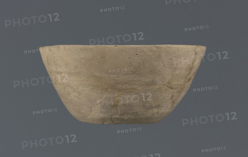 Bowl, unknown Northern Mesopotamian workshop, Early Jazira III period ca. 2600–ca. 2350 BC / Early Dynastic III period ca. 2600–ca. 2350 BC. Clay table vessel; bowl with a flat round bottom with traces of trimming; straight, flaring conical walls terminating in a straight, non-distinctive rim. The whole is asymmetrical. c. 2600–c. 2350 BC; Tell Jassa el Gharbi North-Eastern Syria, Northern Mesopotamia Khabur Basin., A roughly bowl-shaped object with a matte, textured surface sits against a solid background; the object is primarily beige to light brown with areas of darker brown and faint grayish patches, showing subtle horizontal striations and small pits on its surface, and a few visible hairline cracks and irregularities along its rim and lower portion, while the background is a muted charcoal to slate gray.