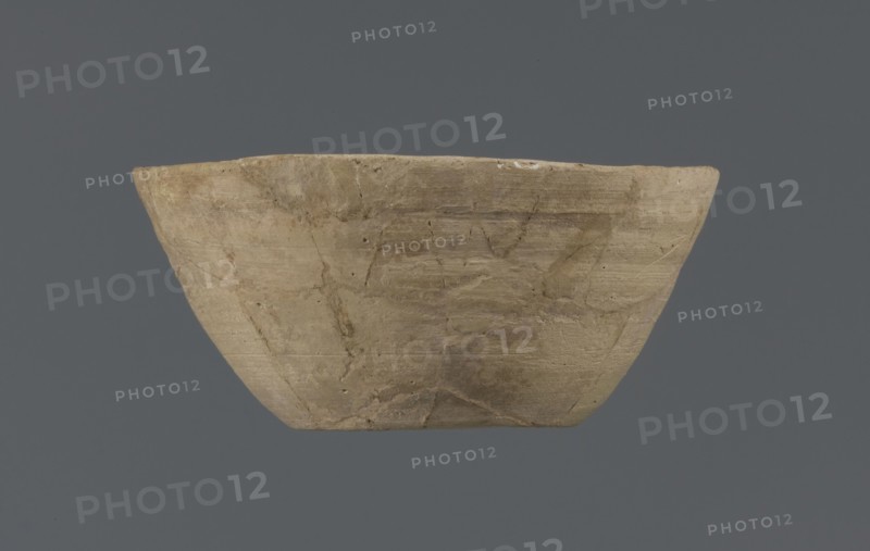 Bowl, unknown Northern Mesopotamian workshop, Early Jazira III period ca. 2600–ca. 2350 BC. Clay table vessel — bowl with a flat round bottom with traces of trimming; straight, flaring conical walls terminating in a straight, non-distinctive rim. The whole is asymmetrical. c. 2600–c. 2350 BC; Early Jazira III period. Tell Jassa el Gharbi North-Eastern Syria; Northern Mesopotamia Khabur Basin., A shallow, wide bowl with an irregular rim displayed against a solid dark gray background, the bowl showing a rough, weathered surface with visible cracks and repair lines; the bowl's color is a mix of beige, light brown, and tan with subtle darker brown streaks and patches, and there are small areas of pale off-white along the rim.