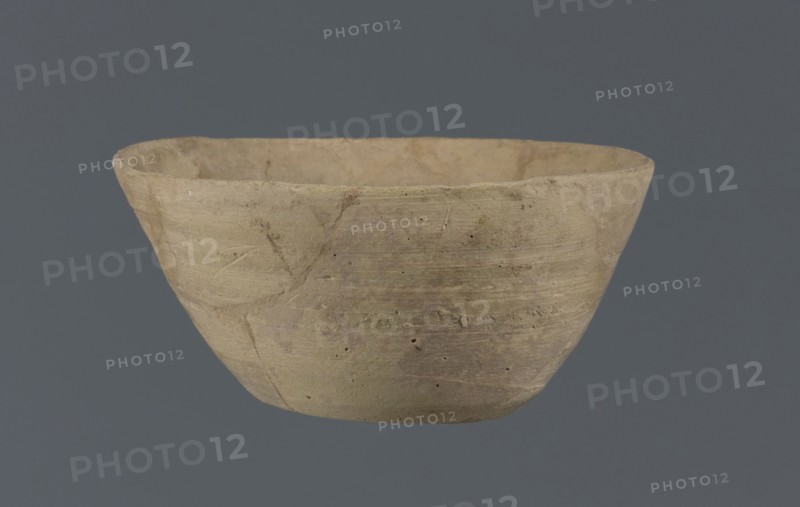 Bowl, unknown Northern Mesopotamian workshop; Early Jazira III period ca. 2600–ca. 2350 BC; Early Dynastic III period ca. 2600–ca. 2350 BC. Clay table vessel bowl with a flat round bottom showing traces of trimming. Straight, flaring conical walls terminating in a straight, non-distinctive rim. The whole is asymmetrical. c. 2600–c. 2350 BC; found at Tell Jassa el Gharbi north-eastern Syria, Northern Mesopotamia Khabur Basin., A roughly rounded, shallow bowl with an uneven rim sits against a flat gray background; the bowl shows a textured, matte surface in shades of beige, tan, and light brown with subtle areas of pale greenish and grayish discoloration, small pits and scratches, and visible horizontal tool marks and faint linear impressions on its exterior.