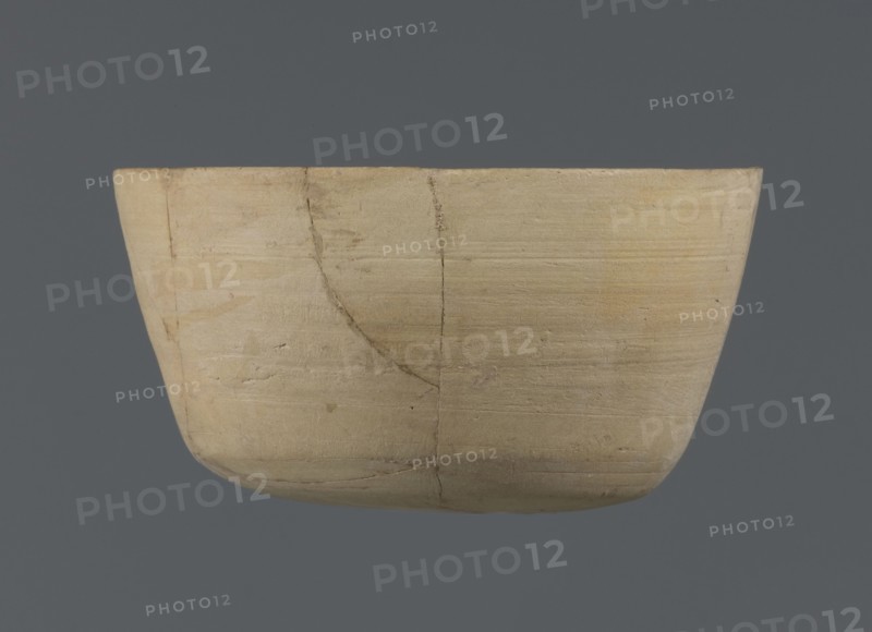 Bowl, unknown northern Mesopotamian workshop; Early Jazira III period ca. 2600–ca. 2350 BC; clay vessel; bowl/table vessel with flat oval bottom from which straight, slightly tilted walls rise in a short arc, terminating in a straight, not distinct rim; Early Dynastic III period ca. 2600–ca. 2350 BC; Al-Jazira Northern Mesopotamia, Khabur basin; found at Tell Jassa el Gharbi north-eastern Syria; height 7 cm, width 12.2 cm., A shallow, rounded container with a smooth, matte surface in pale beige and light tan tones, showing a few darker tan streaks and subtle grayish lines; the vessel has visible horizontal striations and a couple of thin, curved cracks with slightly darker edges; the background is a solid muted slate-gray.