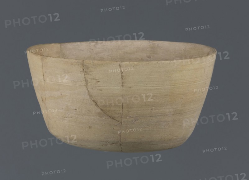 Bowl, unknown northern Mesopotamian workshop, Early Jazira III period ca. 2600–ca. 2350 BC. Clay table vessel — bowl with a flat oval bottom from which straight, slightly tilted walls rise in a short arc, terminating in a straight, not distinct rim. From Tell Jassa el Gharbi North-Eastern Syria, Northern Mesopotamia Khabur basin. Vessel; height 7 cm, width 12.2 cm. Materials and typology: clay, Early Dynastic III period; vice-less vessel; Al-Jazira region., A shallow, rounded bowl with a matte, slightly rough surface displayed against a uniform dark gray background, the bowl showing a light beige to pale tan color with subtle variations and a few darker beige streaks and smudges; the rim is thin and uneven, and the exterior surface bears visible horizontal tool marks and faint cracks where sections meet, while the interior appears a similar pale beige with slight discoloration and texture differences.