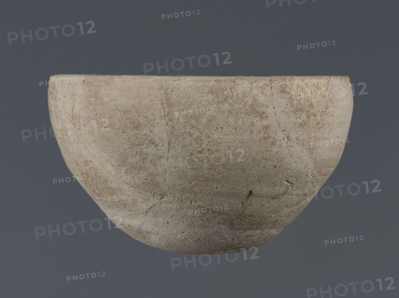 Bowl, unknown Northern Mesopotamian workshop, Early Jazira III period ca. 2600–ca. 2350 BC Early Dynastic III period. Deep clay bowl with a rounded, slightly flattened, carelessly thickened bottom from which the bowl walls emerge in an arched line. In the upper part the walls are slightly inclined toward the center. The straight, low rim is distinguished by a carelessly engraved line on the outside. Origin: Tell Jassa el Gharbi Northeastern Syria, Northern Mesopotamia Khabur Basin. Function: tableware vessel. Dimensions: height 8.7 cm, width 14.4 cm., A single rounded bowl-like object fills most of the frame, showing a rough, weathered surface with visible cracks and pitted texture; the object displays a palette of beige, light tan, pale gray, and hints of muted brown, with subtle variations and mottling across its curved exterior, set against a uniform dark gray background.
