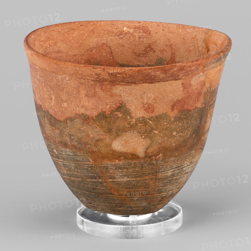 Mug, unknown Northern Mesopotamian workshop. Mug with slightly S-shaped walls and a small flat bottom with a convexity in the center. The rim is thickened in the form of a low, outwardly curled strip. Diagonal scratches are visible on the walls, leaving a technological trace of the vessel being polished. 1st half of the 3rd millennium BC; Early Jazira III period ca. 2600–ca. 2350 BC. Tell Jassa el Gharbi North-Eastern Syria, Northern Mesopotamia Khabur basin. Clay vessel, tableware; height 8.2 cm, width 9.5 cm, weight 156 g., A rounded ceramic vessel resting on a clear circular stand, showing a textured surface with areas of reddish-brown, terracotta, tan, olive green, and darker brown; the upper rim is slightly flared and the interior shares the reddish-brown and tan tones; the exterior displays a mottled pattern with irregular patches and streaks, and the lower portion has horizontal striations and darker brown banding; the clear stand is glossy and colorless, and the background is a...