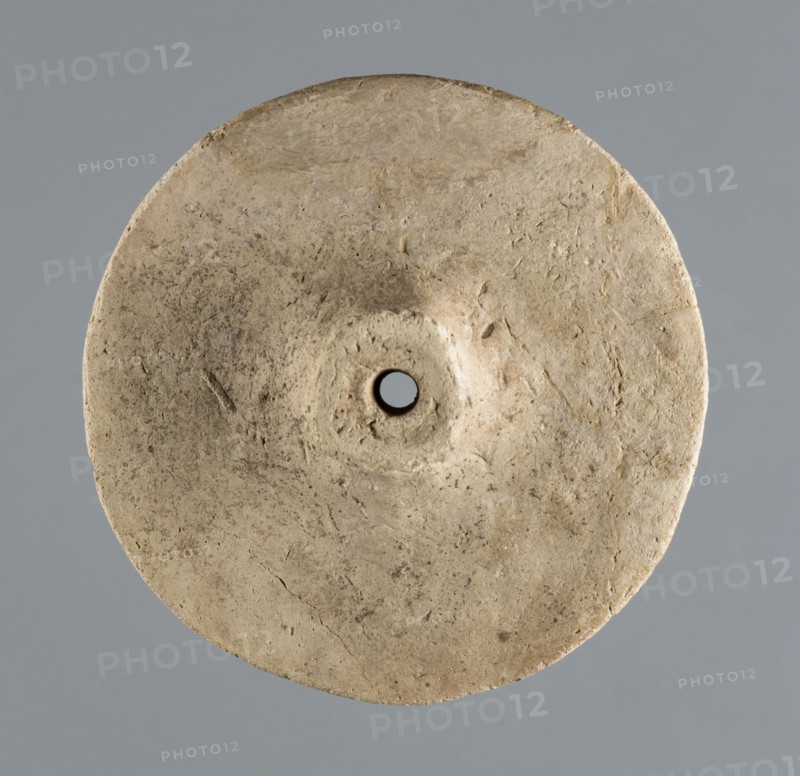 Wheel from a model wagon, unknown northern Mesopotamian workshop, terracotta model wheel with a full disc of lenticular cross-section and a protruding modelled hub on both sides for the axle, c. 2600–c. 2350 BC Early Jazira III / Early Dynastic III period, Northern Mesopotamia Khabur basin, Tell Jassa el Gharbi, Syria, model/toy, height 10 cm, width 3.74 cm, weight 114 g., A roughly circular, flat disc-shaped object with a central round hole, showing a textured surface with irregular marks and small pits; the disc is a beige to light tan color with areas of darker brownish-gray speckling and slight discoloration, set against a smooth medium gray background.