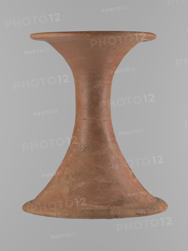 Vessel base. Funnel-shaped vessel base, narrowest at about 2/3 of the height. Both rims are oval in cross-section, slightly upturned. The upper rim has a slightly wavy line. Made of Nile clay. The outer surface and the surface of the upper rim are smoothed, covered with a layer of engobe and polished. The remaining inner surface is smoothed. Vessel color: red. Vessel glued together from several fragments; gaps filled with plaster. The surface is eroded in places. Comparative material: Reisner, A history of Giza necropolis, vol. I, fig. 282, C,4240, ca. 2686–ca. 2181 BC; Old Kingdom, Edfu, vessel. Height 23.9 cm; diameter 16.9 cm rim; diameter 19.6 cm bottom., A single hourglass-shaped terracotta object with a flared top and base, showing a textured surface with irregular patches and spots; colors visible include shades of reddish-brown, earthy orange, and small areas of darker brown, set against a neutral light gray background.