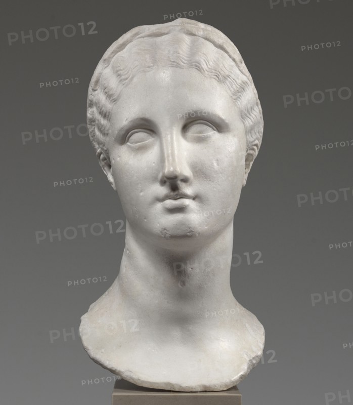 Head of a woman Berenice II or Arsinoe II, unknown creator; Greek sculpture, female portrait, Hellenistic period, 330–30 BC; Alexandria; sculpture; restitution Przeworsk, A white marble bust of a woman's head and neck is shown against a neutral gray background; the sculpture features wavy hair pulled back with a headband, smooth facial features with subtle relief around the eyes and mouth, and a gently rounded chin and long neck, with the lower edge of the bust rough and uneven resting on a small rectangular base; visible colors include white, off-white, gray, and beige.