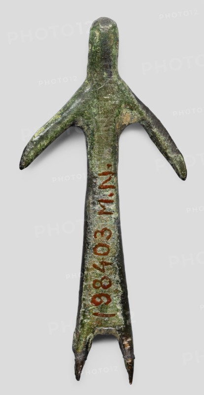 Votive figurine of a woman, votive offering, bronze female figurine, 1st half of the 6th century BC, Etruria; height 6.1 cm, width 2.9 cm, depth 0.5 cm., A weathered metal object shaped like a stylized human figure with an elongated body and a rounded head, showing a greenish patina with areas of darker brown and lighter green; it has a pair of outstretched arm-like projections and a split lower end forming two pointed prongs, with traces of reddish-brown markings along the central shaft.