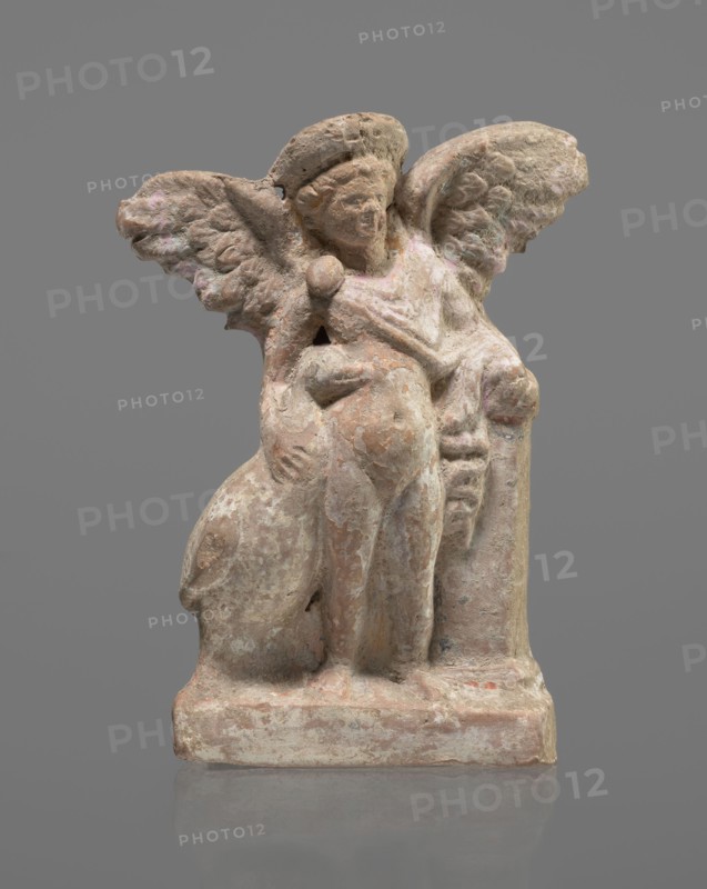 Terracotta figurine of Eros with a goose, terracotta, Eros mythology, Greek mythology, figurines from a mold. Figurine of a standing Eros. The figure, positioned frontally, slightly leaning back and facing left, stands on a low, rectangular base. On the left, he leans against a small column, and on the right, he holds a goose standing nearby by the neck. The left hand, clasped at the elbow, rests on the top of the column, holding a short cloak halfway up. The cloak, fastened with a fibula over the right shoulder, covers the chest and forms folds and flows along the small column. The hair, arranged in curls, is covered in the upper part by a high diadem. The outstretched wings, with straight, defined feathers, are slightly tilted back. Scratches faded, traces of polychrome. 2nd half of the 1st century BC, Asia Minor. Figurine, height 14 cm, width 10.2 cm, depth 3.4 cm, weight 0.18 kg., A weathered small sculpture of a winged figure carved from a light brownish-pink stone, showing a stan...