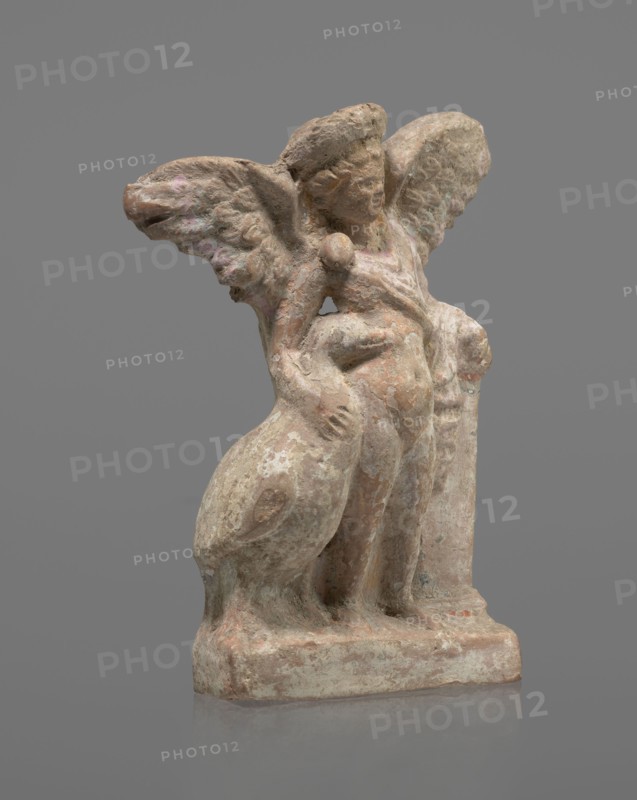 Terracotta figurine of Eros with a goose. Figurine of a standing Eros, positioned frontally, slightly leaning back and facing left, standing on a low rectangular base. He leans on a small column at the left and holds a goose by the neck at the right. The left hand, clasped at the elbow, rests on the top of the column and holds a short cloak halfway up. The cloak, fastened with a fibula over the right shoulder, covers the chest and forms folds and flows along the column. Hair arranged in curls and covered in the upper part by a high diadem. Outstretched wings with straight, defined feathers slightly tilted back. Scratches faded, traces of polychrome. Terracotta; figurine from a mold. 2nd half of the 1st century BC, Asia Minor. Height 14 cm, width 10.2 cm, depth 3.4 cm, weight 0.18 kg., A weathered stone figurine with a rough surface showing tones of beige, pale pink, muted green, and gray stands on a rectangular base; the figure has a rounded head with indistinct facial features, a pair...