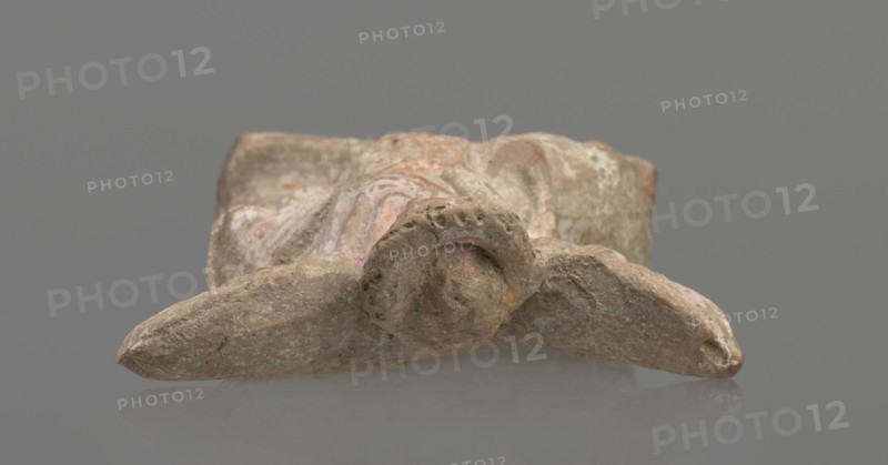 Terracotta figurine of Eros with a goose. Figurine of a standing Eros: the figure, positioned frontally, slightly leaning back and facing left, stands on a low rectangular base. On the left he leans against a small column; on the right he holds a goose by the neck. The left hand, clasped at the elbow, rests on top of the column holding a short cloak halfway up. The cloak, fastened with a fibula over the right shoulder, covers the chest and forms folds and flows along the small column. Hair arranged in curls, covered in the upper part by a high diadem. Outstretched wings with straight, defined feathers are slightly tilted back. Scratches faded; traces of polychromy. Terracotta, from Asia Minor; 2nd half of the 1st century BC. Figurine from a mold. Height 14 cm, width 10.2 cm, depth 3.4 cm, weight 0.18 kg., A weathered stone or clay object rests on a smooth gray surface against a neutral gray background, showing a rough, textured surface with irregular shapes and worn edges; the object h...