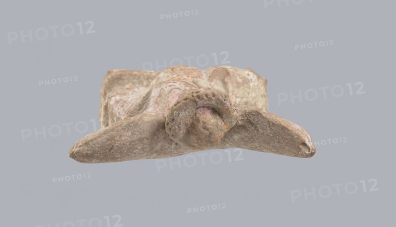 Terracotta figurine of Eros with a goose. Terracotta; figurine from a mold; 2nd half of the 1st century BC; Asia Minor. Figurine of a standing Eros positioned frontally, slightly leaning back and facing left, standing on a low rectangular base. On the left he leans against a small column; on the right he holds a goose by the neck. The left hand, clasped at the elbow, rests on the top of the column and holds a short cloak halfway up. The cloak, fastened with a fibula over the right shoulder, covers the chest and forms folds and flows along the small column. Hair arranged in curls, covered in the upper part by a high diadem. Outstretched wings with straight, defined feathers, slightly tilted back. Scratches faded, traces of polychrome. Height 14 cm; width 10.2 cm; depth 3.4 cm; weight 0.18 kg., A weathered stone or clay object with an irregular, rounded central form and a curved protrusion extending outward, showing a rough textured surface with patches of pale pink, light brown, beige, ...