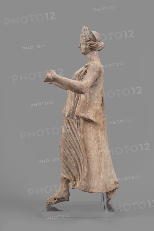Figurine of the goddess Nike, Nike mythology, Greek deity, female figurine, 1st half of the 2nd century BC, Myrina site of finding, figurine, height 27 cm, width 12 cm, depth 11 cm, weight 0.47 kg, A weathered statue of a robed female figure shown in profile, made of a light beige to pale brown stone with areas of darker brown patina and faint traces of reddish tones, standing on a gray rectangular base with a metal support visible; she faces left with her hair pulled back under a cap, wearing a draped garment that shows carved folds and pleats cascading down the front and side, arms extended forward with hands clasped together, bare feet stepping forward with visible toes, and the surface shows rough texture and small abrasions.