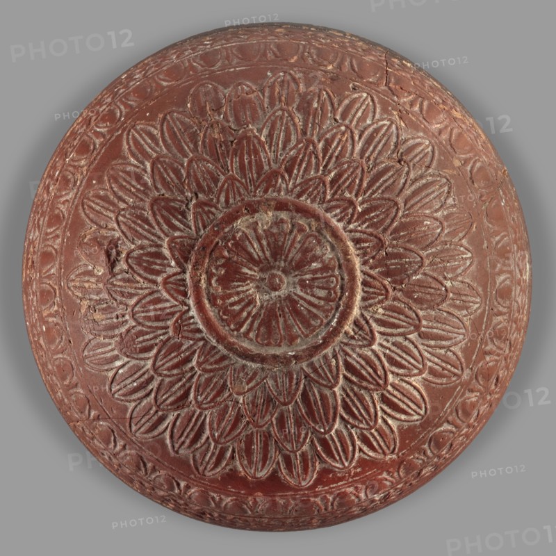 Megarian bowl, Monogramist's Workshop, 2nd half of the 2nd century BC, Ephesus, Ionia; vessel; weight 0.134 kg, A circular carved object with a raised floral medallion design occupying the center, showing layered petal shapes radiating outward and a round central rosette, all rendered in a reddish-brown color with lighter worn highlights revealing a paler tone, set against a neutral gray background; the surface shows carved linear and scalloped border patterns around the rim and areas of surface wear and small chips.