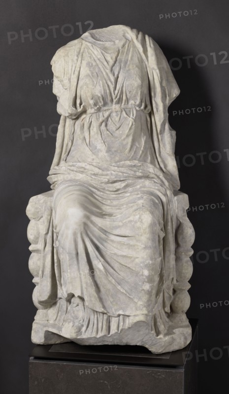 Statue of the goddess Cybele on a throne, Cybele myth., statue of a deity, Roman sculpture, 1st century BC–1st century AD, Asia Minor, sculpture; height 84 cm, width 43 cm, depth 39 cm, weight 181 kg, A headless stone statue of a seated draped figure sits on a blocky base against a dark background, showing folded and gathered garments with detailed carved folds and flowing drapery in pale grey and off-white tones, with areas of subtle discoloration and weathering; the figure's arms are partially present beneath the cloth, and the seat has rounded carved elements visible at the sides, while the base beneath the statue is a darker stone with a smooth surface in charcoal and deep grey colors.