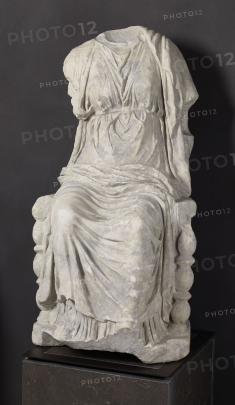 Statue of the goddess Cybele on a throne, Puslowski, Scen.Gal.Klass., Cybele myth., statues of deities, Roman sculpture, 1st century BC–1st century AD, Asia Minor, sculpture; height 84 cm, width 43 cm, depth 39 cm, weight 181 kg, A headless and armless white stone statue of a seated figure draped in layered flowing garments with detailed folds, resting on a carved chair with rounded vertical elements on the sides, displayed against a dark gray background on a dark pedestal; visible colors include white and off-white tones of the statue, dark gray of the backdrop, and black and charcoal of the pedestal.