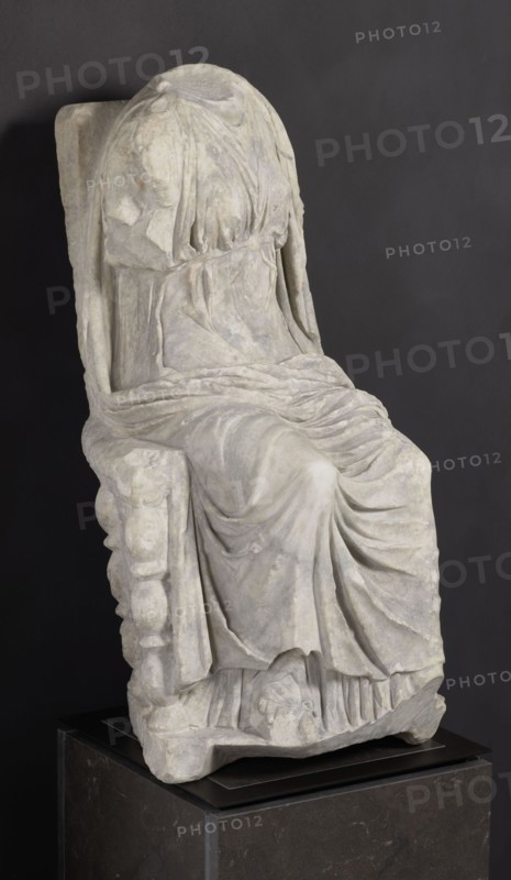 Statue of the goddess Cybele on a throne; creator, Cybele myth., statues of deities; Roman sculpture, 1st century BC–1st century AD; Asia Minor possibly; sculpture; height 84 cm, width 43 cm, depth 39 cm, weight 181 kg., A draped seated stone figure on a pedestal, shown against a dark gray background, with the drapery forming flowing folds and pleats over the torso, arms, and legs; the head is covered and lacks facial details, and the figure’s garments show textured surfaces and soft curves; the seat has a sculpted side with rounded protrusions and the base rests on a dark rectangular pedestal. Colors visible include shades of light gray and off-white on the sculpture, dark gray on the background, and darker gray and black tones on the pedestal.
