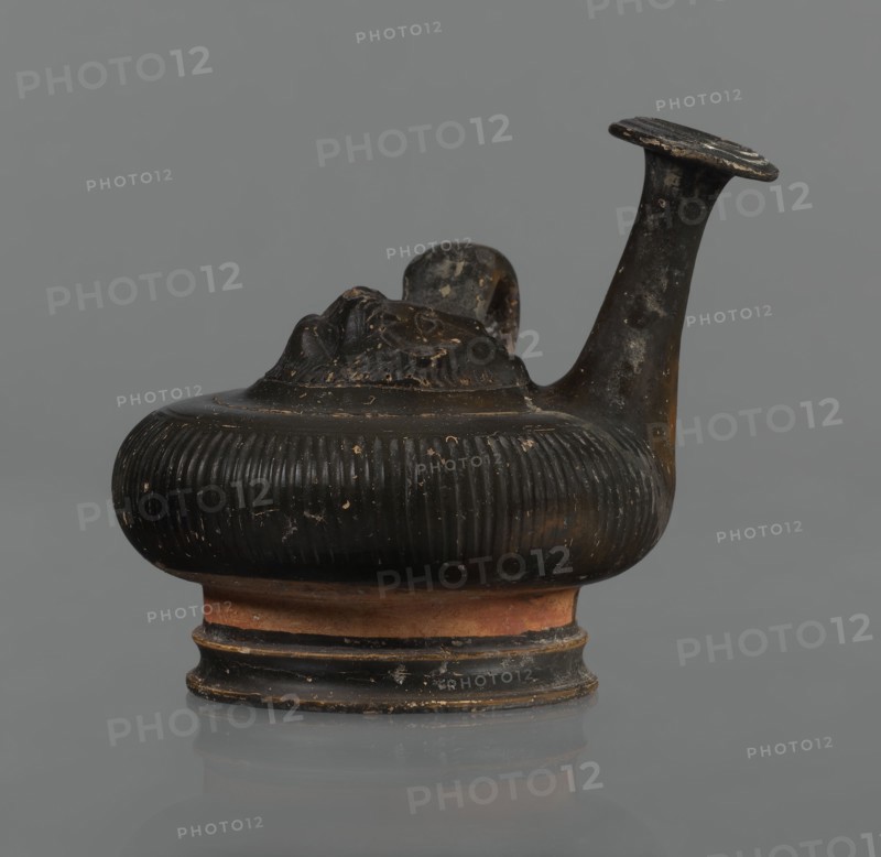 Black-finned guttus, guttus, Greek pottery, black-finned pottery. Guttus with a high foot composed of a profiled lower part and a cylindrical, narrower upper part this part is not covered with the finis. Body in the shape of a flattened sphere with vertical grooves. A band-shaped handle on the right side of the vessel. Long neck with a wide, flared rim. On the upper surface is a convex medallion, surrounded by a single band, depicting the head of Heracles in a lion's skin. Round face, massive nose, wide mouth, large almond-shaped eyes. The lion skin and mane frame Heracles' face. Finish ranges from black to shiny brown. Condition: the handle and neck are cracked and glued; cracks and abrasions in the finish. 3rd century BC, Calvi place of manufacture, vessel, guttus; height 10.7 cm, diameter 11.5 cm., A small sculpted vessel with a bulbous, ribbed body and a short flared base, shown in profile against a plain grey background; the vessel is predominantly dark brown to black with worn ar...