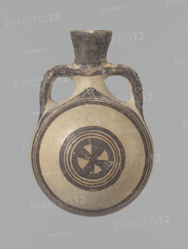 Canteen, Cyprus. Canteen decorated with concentric circles. Vessel with a spherical, slightly flattened body width on one side 8.5 cm, on the other 7.3 cm and two vertical, cylindrical vises and a narrow neck. Slightly flaring spout reconstructed. Pinkish clay. Creamy engobe, black-brown decoration. On the handles, horizontal lines framed on the sides by thick lines, which are connected at the neck at the attachment point, while below the vises they intersect below the handles. The two flattened sides of the body are decorated identically. Two sets of concentric circles, composed of four lines each: two narrow lines framed on both sides by wider lines. The outer line of the outer band overlaps the neck of the vessel and slightly extends over the vice mounts, widening in these places. The center of the inner band is filled with a Maltese cross motif. Good condition: reconstructed spout and upper half of the neck. Limescale deposits have been removed from the surface of the vessel. Numer...