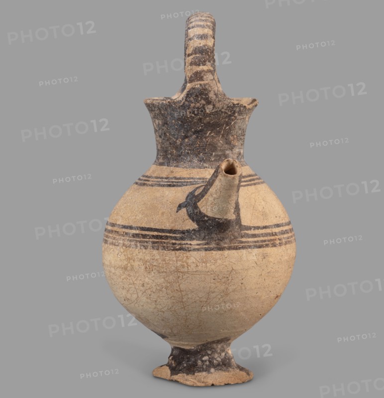 Jug, Jug on a ring-shaped foot from which a low, wide stem extends, transitioning into a belly. The upper part of the spherical body has a narrow spout. The wide, flaring neck terminates in a simple spout, from both sides of which a vice shaped like a flattened cylinder extends above the vessel. Decoration in brown varnish: concentric circles around the vessel four below and three above the spout. The foot and stem are varnished on top; the neck and outer side edges of the vice are varnished. The upper part of the vice is decorated with a row of transverse stripes. A strip of varnish encircles the spout at the base, and three stripes decorate the spout on the sides and top. Dated 1050–950 BC; Cyprus, Geometric Period I District IA. Vessel. Height 12.8 cm; width 6.5 cm., A rounded ceramic vessel with a narrow neck, a flared lip, a single curved handle arching from the lip to the neck, and a short spout protruding from the shoulder; the body sits on a small foot. The surface shows a beig...
