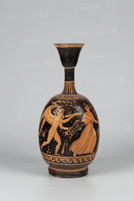 Red-figure aryballus lekythos depicting Eros and a woman, attributed to the Darius Painter active 340–320 BC, Apulia, 340–320 BC; vessel lekythos, aryballus lekythos, height 23.3 cm, width 10.7 cm., A rounded ceramic vessel with a narrow neck and flared mouth sits against a light gray background; the vessel's surface shows a palette of black and orange with areas of worn brown. The body features an orange-figured scene on a black field depicting a winged figure reaching toward a draped standing figure, both rendered in orange with black detailing; decorative bands and a wave-like border encircle the base in orange and black, and the neck shows alternating vertical orange and black stripes leading to a largely black flared rim that has worn orange edges.