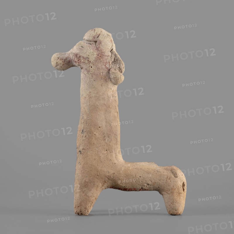 Horse figurine, unknown Cypriot workshop. Schematic horse figurine. Plastically modeled muzzle, strongly rounded at the tip. Eyes marked with convexities. Mane attached. Neck strongly elongated in relation to the rest of the body, flattened at the rear edge, imitating a mane. Body narrow in relation to the neck, cylindrical. Front legs spread out to the sides, also cylindrical. Hind legs designed as a single unit in the form of a cylinder. Traces of polychrome visible on the surface: muzzle and mane reddish-brown, eyes highlighted in black. Traces of black paint also visible on the neck and chest of the animal. 750–600 BC Cypro-Archaic period II, Cyprus. Terracotta figurine, height 10.8 cm, width 7.8 cm., A clay figurine shaped like an abstract animal or creature stands against a gray background; its surface is rough and textured with visible pitting and small holes, and its overall color is a pale beige with patches of light brown and faint reddish areas, especially near the head and ...