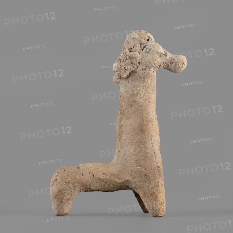 Horse figurine, unknown Cypriot workshop. Schematic horse figurine. Plastically modeled muzzle, strongly rounded at the tip. Eyes marked with convexities. Mane attached. Neck strongly elongated in relation to the rest of the body, flattened at the rear edge, imitating a mane. Body narrow in relation to the neck, cylindrical. Front legs spread out to the sides, also cylindrical. Hind legs designed as a single unit in the form of a cylinder. Traces of polychrome visible on the surface: muzzle and mane reddish-brown, eyes highlighted in black. Traces of black paint also visible on the neck and chest of the animal. 750–600 BC previously dated 6th century BC; Cypro-Archaic II, Cyprus, terracotta figurine, height 10.8 cm, width 7.8 cm., A weathered clay sculpture of an animal-like form stands against a plain gray background, showing a tall vertical body with a horizontal limb at the base creating an L-shaped silhouette; the surface is rough and pitted with irregular texture and small holes, ...