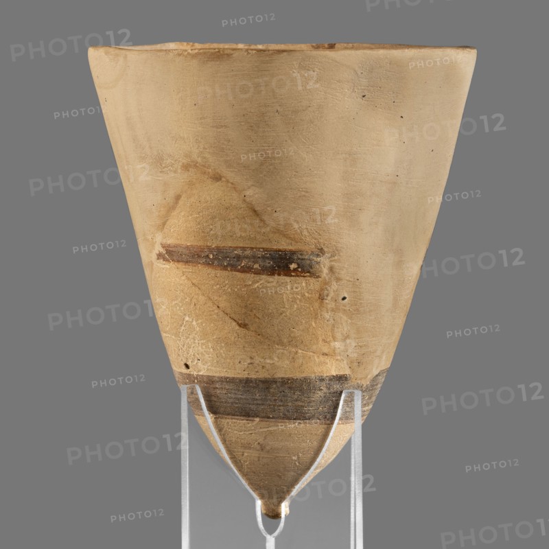 Cup, unknown Middle Assyrian workshop; Mitannian painted pottery Northern Mesopotamia, Mitanni Kingdom in Northern Mesopotamia; Eastern Gallic Scenography; 14th–13th century BC, Middle Assyrian period; vessel., A conical, beige ceramic vessel with a pointed bottom sits on a clear support against a gray background, showing a worn surface texture with scratches and small pits, a few horizontal dark brown bands and a faint darker area on one side, and subtle variations of tan and ochre tones across the body.