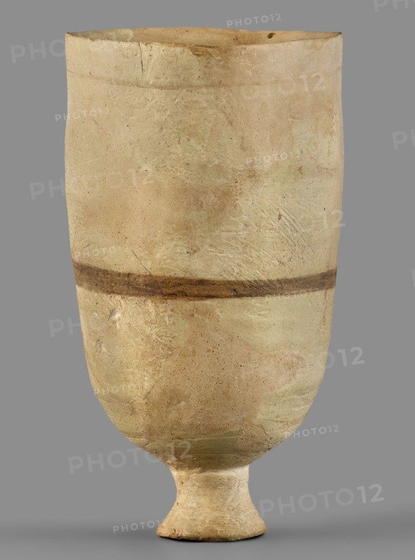 Cup, unknown Mitannian workshop, Mitannian pottery 15th–14th century BC, stemmed cup, Mitanni, Northern Mesopotamia Kingdom of Mitanni, material: pottery, function: vessel, period: Mitannian 15th–14th century BC, An upright ceramic vessel with a rounded body that tapers to a short pedestal foot, showing a matte, weathered surface in shades of beige, cream, light tan, and pale green, with a darker brown horizontal band around the midsection and scattered small darker specks and patches; the rim is slightly irregular with minor chips and the surface displays subtle texture variations, scratches, and areas of wear.