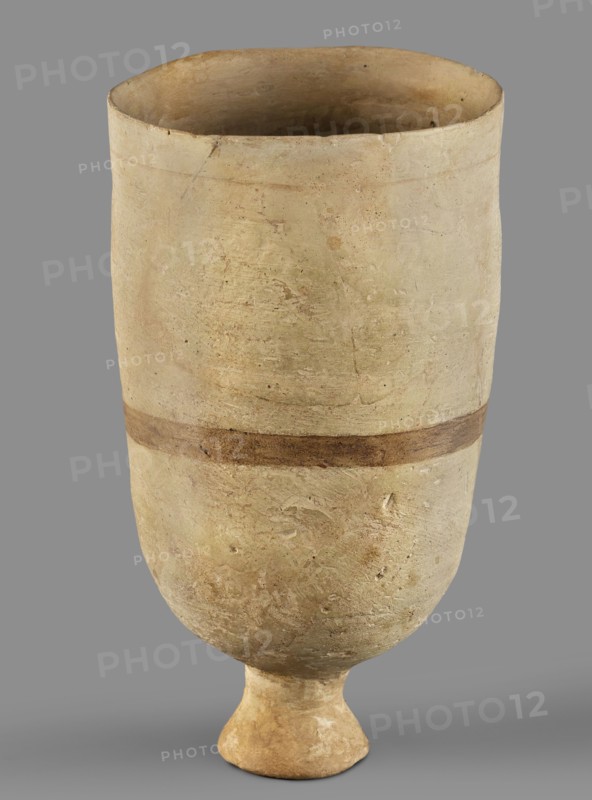 Cup, unknown Mitannian workshop, Mitannian pottery 15th–14th century BC, stemmed cup, material culture, Mitanni, Near Eastern pottery, Eastern Gallic scene, 15th–14th century BC; Mitannian period, Northern Mesopotamia Kingdom of Mitanni, vessel, A tall, cylindrical ceramic vessel with a rounded base that tapers to a short pedestal foot, shown against a plain gray background; the vessel surface is textured and worn with an overall pale beige or sandy color, a slightly darker band of brown encircles the midsection, and there are subtle variations in tone including light tan and off-white patches as well as small areas of darker brown and faint greenish discoloration.