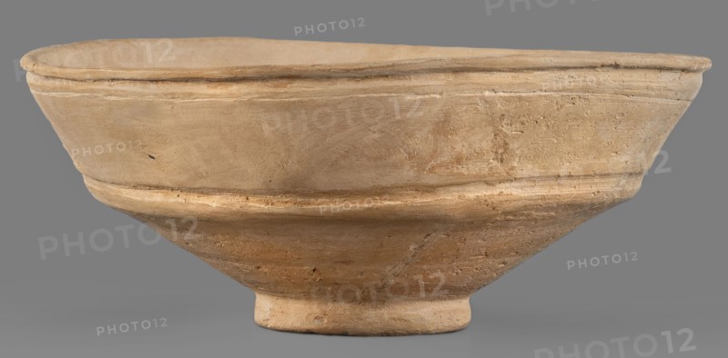 Bowl, unknown Middle Assyrian workshop; Middle Assyrian pottery, bowls on a ring-shaped foot; bowl with broken profile; vertical wall above the break slightly inclined and decorated with two grooves; ring-shaped foot; 13th–12th century BC; Middle Assyrian period, Northern Mesopotamia; vessel., A shallow, wide bowl with a rounded rim and a small circular foot, shown against a plain background; the bowl surface has a textured, slightly rough appearance with visible tool marks and irregularities, and a raised band running around its midsection; colors visible include beige, tan, light brown, and hints of darker brown in weathered areas.