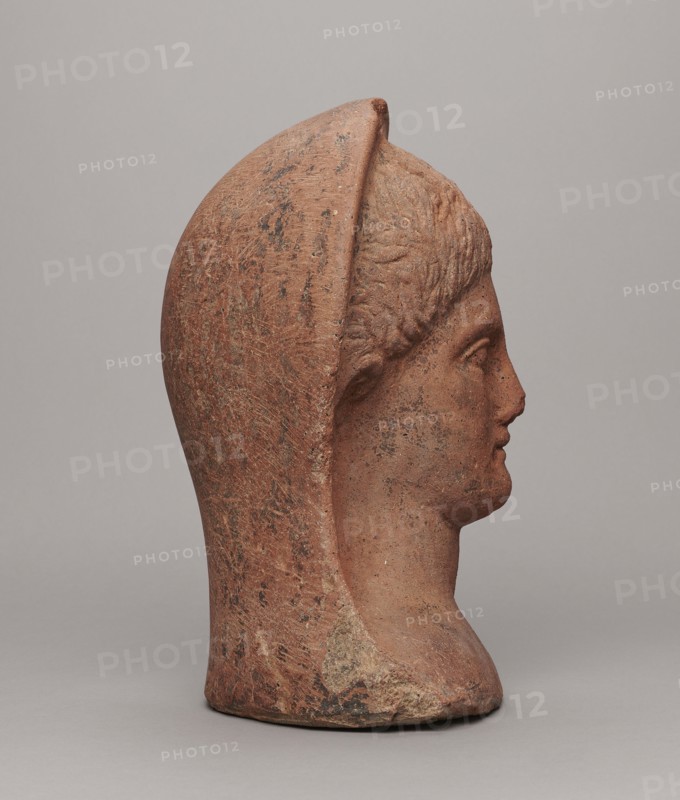 Votive head, unknown creator; Hellenistic period, 332–30 BC; Central Italy; sculpture; height 30 cm, width 22.5 cm, depth 17.5 cm, weight 6.1 kg., A terracotta-colored sculpted head in profile facing right, with a rounded helmet-like covering that extends down the back and sides, showing textured surface marks and patches of darker brown and black discoloration, a smooth forehead and stylized short hair texture, a defined nose, lips, and chin, and a similarly colored neck and base resting on a neutral pale gray surface with a pale gray background.