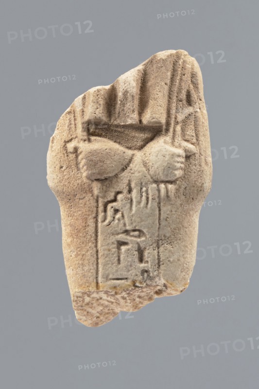 Ushabti - fragment, Mummy-shaped figurine; flat pillar on the back. Preserved central fragment from shoulder right arm damaged to hip height. Arms crossed on the chest, right folded over left; only hands visible from under the shroud. Hoes in both hands with slightly curled ends. In the right hand, a smooth rope bearing a trapezoidal basket with a semicircular handle slung over the left shoulder. Convex tools; engraved basket. Remains of a three-part smooth wig; at the back the wig is slightly wider than the pilaster. Fragmentarily preserved straight, long beard. Engraved inscription placed in a single column on the front of the legs, text bounded by two vertical lines. Excavated at Tell Atrib, shabti; height 6 cm, width 3.4 cm., A weathered beige stone fragment with a textured, grainy surface sits against a flat gray background; the fragment shows carved, rounded forms resembling a pair of clasped hands or rounded shapes near the top with vertical grooves and ridges above them, and a ...