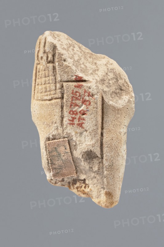 Ushabti - fragment, Mummy-shaped figurine, funerary offering. Flat pillar on the back. Preserved central fragment of the figurine—from the shoulder right arm damaged to hip height. Arms crossed on the chest, right folded over the left. Only the hands visible from under the shroud. Hoes in both hands; ends slightly curled. In the right hand a smooth rope supporting a trapezoidal basket with a semicircular handle slung over the left shoulder. Convex tools; engraved basket. Remains of a three-part smooth wig; at the back the wig is slightly wider than the pilaster. Fragmentarily preserved beard; originally straight and long. Engraved inscription placed in a single column on the front of the legs, text bounded by two vertical lines. Findspot: Tell Atrib, shabti. Dimensions: height 6 cm, width 3.4 cm., A fragment of a light beige stone with a rough, pitted surface suspended against a muted gray background, showing carved linear grooves and shallow rectangular impressions on one face, traces...