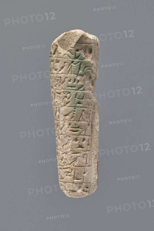 Ushabti - fragment. Mummy-shaped figurine fragment of the legs preserved from hips to ankles; the back rests on a pillar. Inscribed hieroglyphs in ten horizontal bands around the legs, separated by thin lines. Function: ushabti; grave good. Materials: probably faience/stone not specified. Date: 332–30 BC, Ptolemaic Period. Place of discovery: Tell Atrib. Dimensions: height 8.5 cm, width 3.8 cm., A narrow, elongated stone object with a rounded top and bottom is shown against a plain gray background; the stone is light beige to sandy in color with areas of pale greenish discoloration, and its surface features several carved rectangular panels and shallow relief shapes arranged in vertical rows, with some edges chipped and worn, giving an overall weathered, pitted texture.